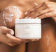 A woman holding a jar and exfoliating her body with FarmHouse Fresh Sweet Cream body scrub that leaves skin soft and smelling amazing.