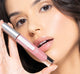 A woman holding FarmHouse Fresh Vitamin Glaze Oil Infused Lip Gloss in Delicate Rose color by her face, demonstrating the rich color of the lip gloss and her soft, well moisturized lips.