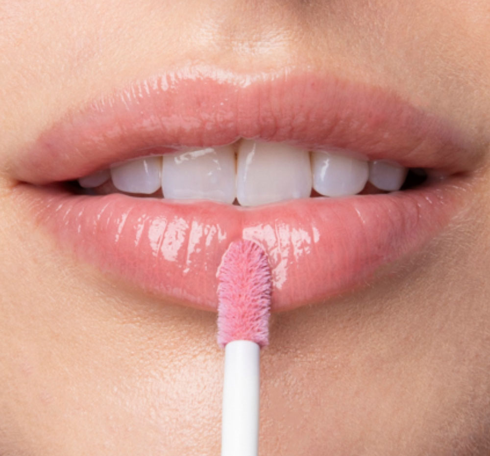 A woman is applying FarmHouse Fresh Vitamin Glaze Lip Gloss in Delicate Rose color that hydrates to lips.