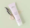 A tube of Farmhouse Fresh Buttermilk Lavender Steeped Milk Lotion® for hands with a creamy texture smudge and a twig of lavender flowers on a green background.