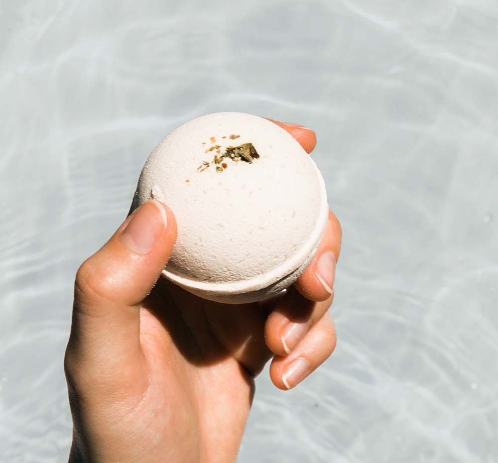 A hand holding FarmHouse Fresh Rosemary-Mint Hemp-Infused Large Fizzing Bath Soak with Swellness Fortune above bath water.