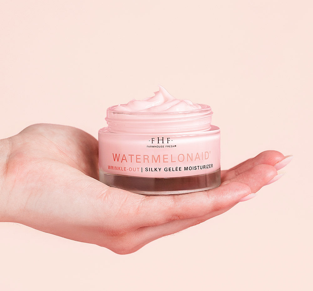 Hand holding a jar of Farmhouse Fresh Watermelonaid cream infused with watermelon extract and CBD on a pink background.