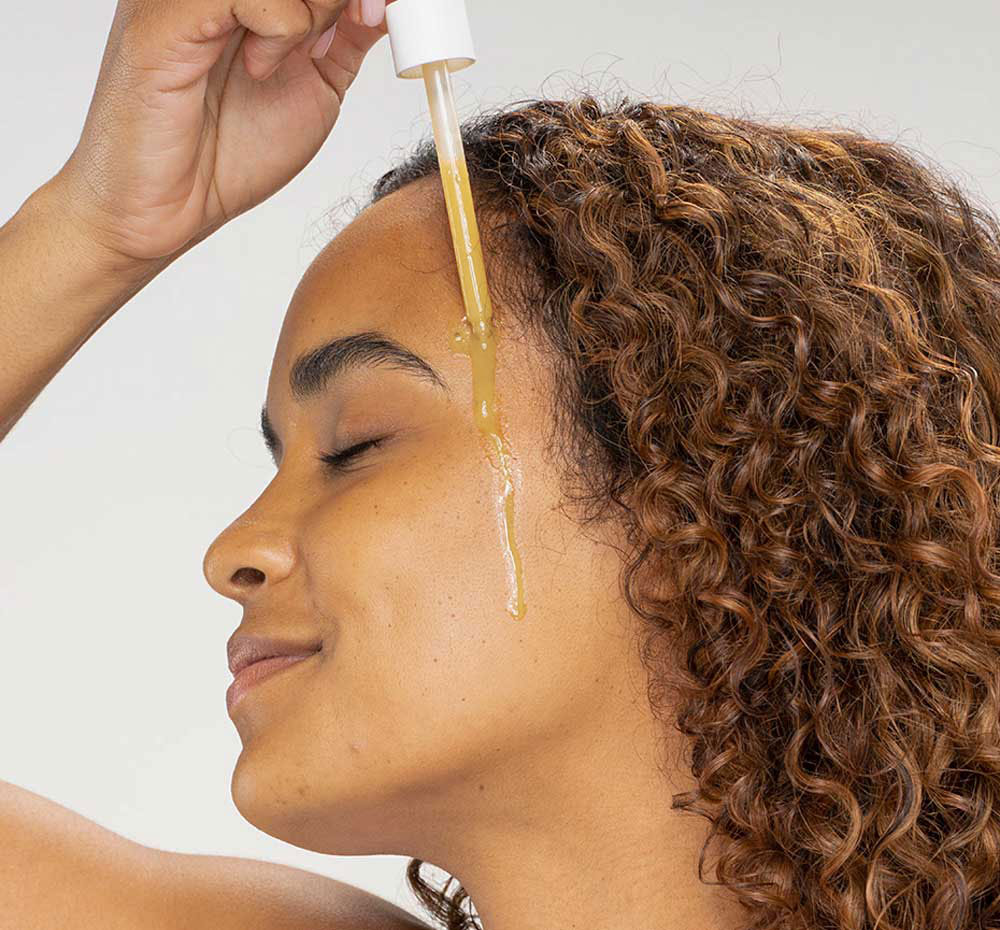 A woman is applying FarmHouse Fresh Bakuchiol Booster serum on her face to brighten and soother her skin.