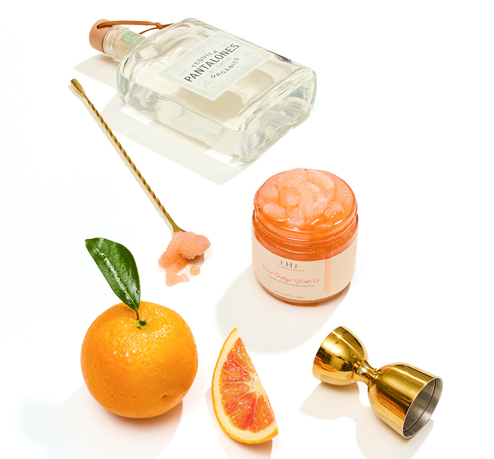 Blood Orange Glowrita body polish with Pantalones Blanco tequila bottle, whole orange, orange slice, and gold bar spoon