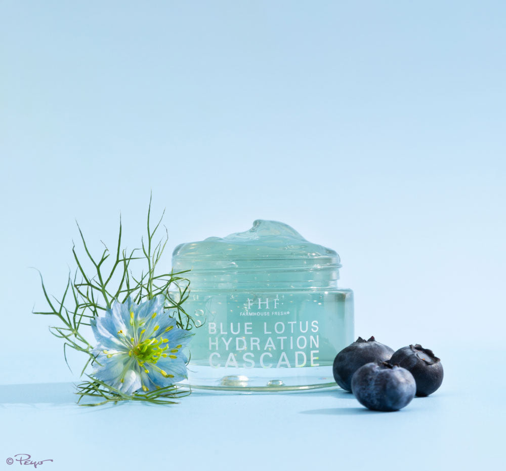 A jar of FarmHouse Fresh Blue Lotus Hydration Cascade Moisturizer next to blue lotus flower and honeyberries, the key natural ingredients of this face lotion that help calm, smooth and visibly brighten the look of skin.