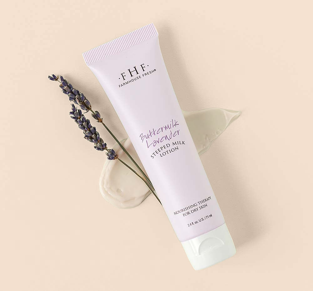 A tube of Farmhouse Fresh Buttermilk Lavender Steeped Milk Lotion® for hands with a creamy texture smudge and a twig of lavender flowers on a green background.