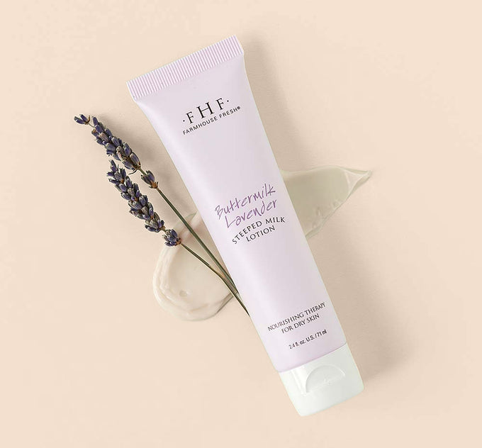 A tube of Farmhouse Fresh Buttermilk Lavender Steeped Milk Lotion® for hands with a creamy texture smudge and a twig of lavender flowers on a green background.