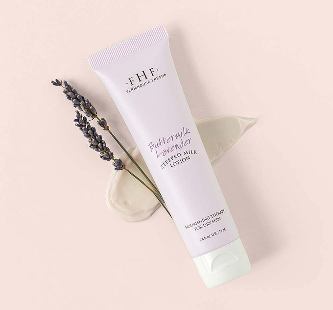 A tube of Farmhouse Fresh Buttermilk Lavender Steeped Milk Lotion® for hands with a creamy texture smudge and a twig of lavender flowers on a green background.