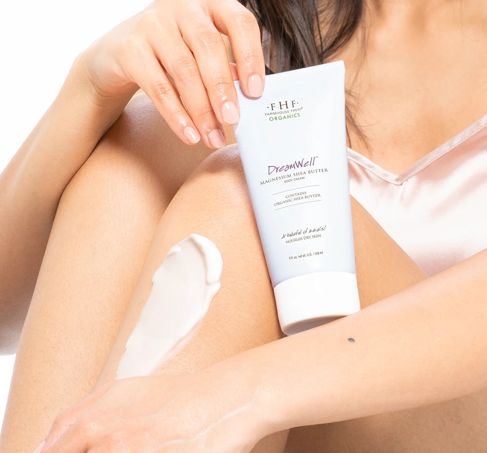 Woman sitting and holding a tube of DreamWell™ Magnesium Organic Shea Butter Body Cream applying the product to their leg.