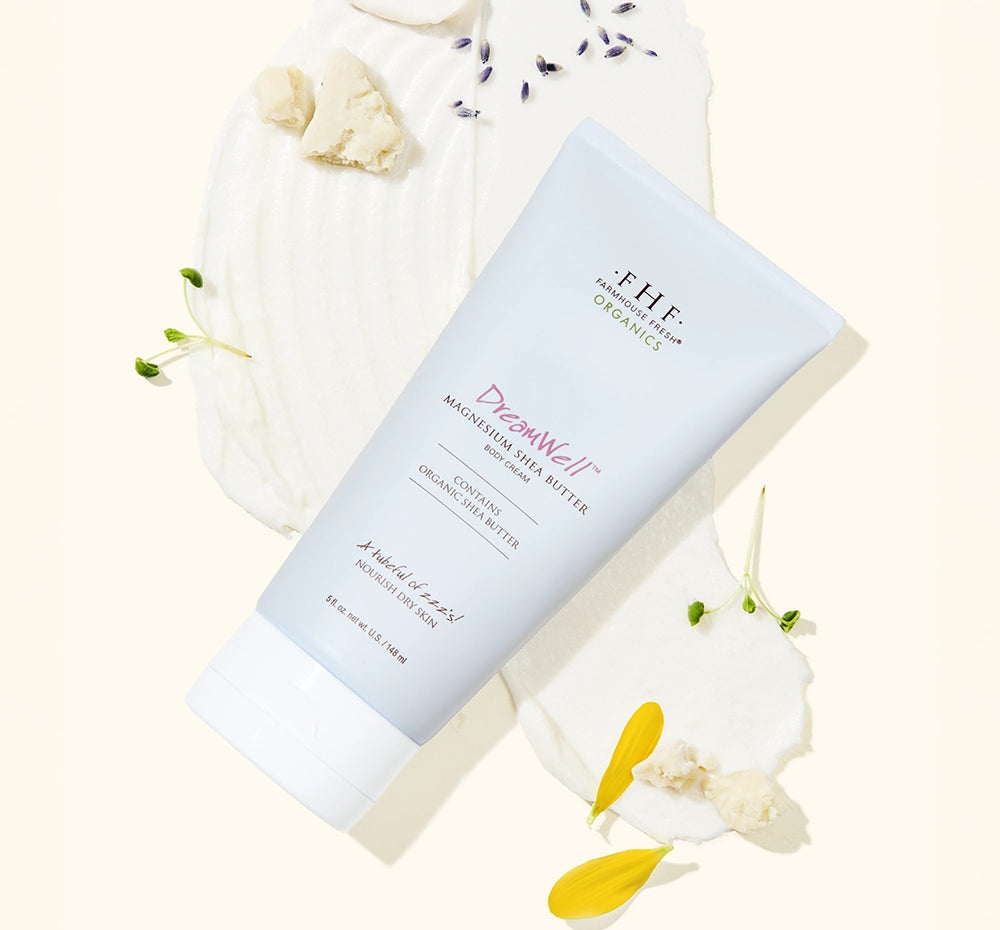 Tube of DreamWell™ Magnesium Organic Shea Butter Body Cream laying on a product swipe with ingredients sprinkled around.