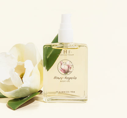 Bottle of Honey-Magnolia body oil with a Magnolia flower sitting next to the bottle, on a light cream background.