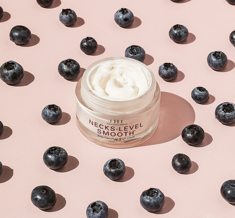 A jar of FarmHouse Fresh Necks-Level Smooth Neck Cream surrounded by bilberries, one of the main natural ingredients with calming and redness-reducing properties.