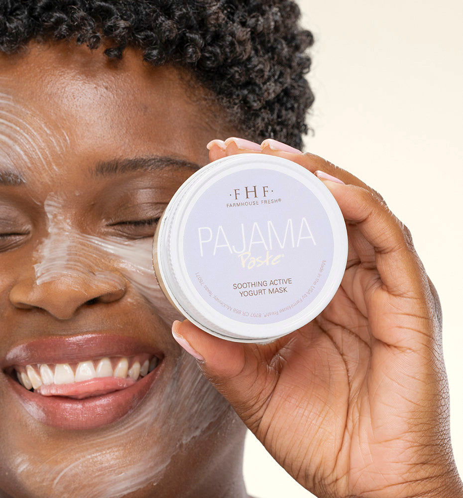 A woman with Pajama Paste Soothing Active Yogurt Mask by FarmHouse Fresh on her face and a jar in her hand, smiling while enjoying the pleasant honey scent of the product.