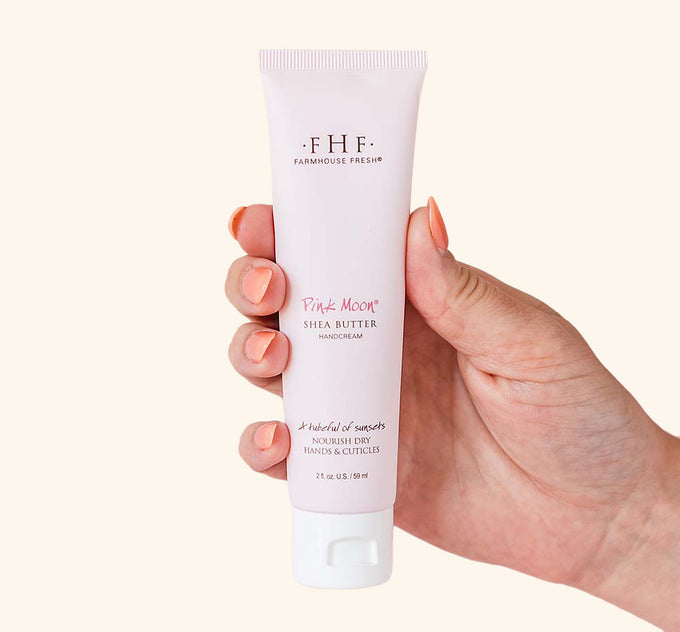 A hand holding a tube of FarmHouse Fresh Pink Moon Shea Butter hand cream that leaves skin hydrated and soft.