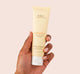 A hand is holding a tube of Farmhouse Fresh Rainbow Road Shea Butter hand lotion for dry skin.