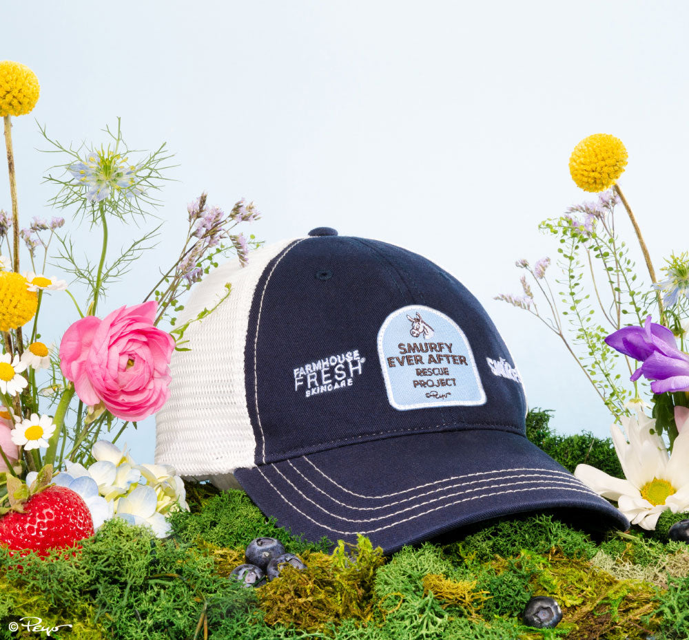 FarmHouse Fresh Smurfin' Cool Trucker Hat surrounded by flowers. All profits are donated to animal rescue initiatives around the country.