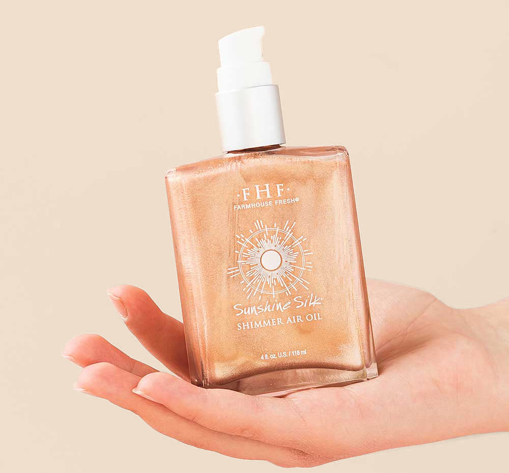 A hand holding a bottle of FarmHouse Fresh Sunshine Silk Shimmer Air Oil made with natural ingredients that moisturize and soften skin while giving it a rose-gold glow.