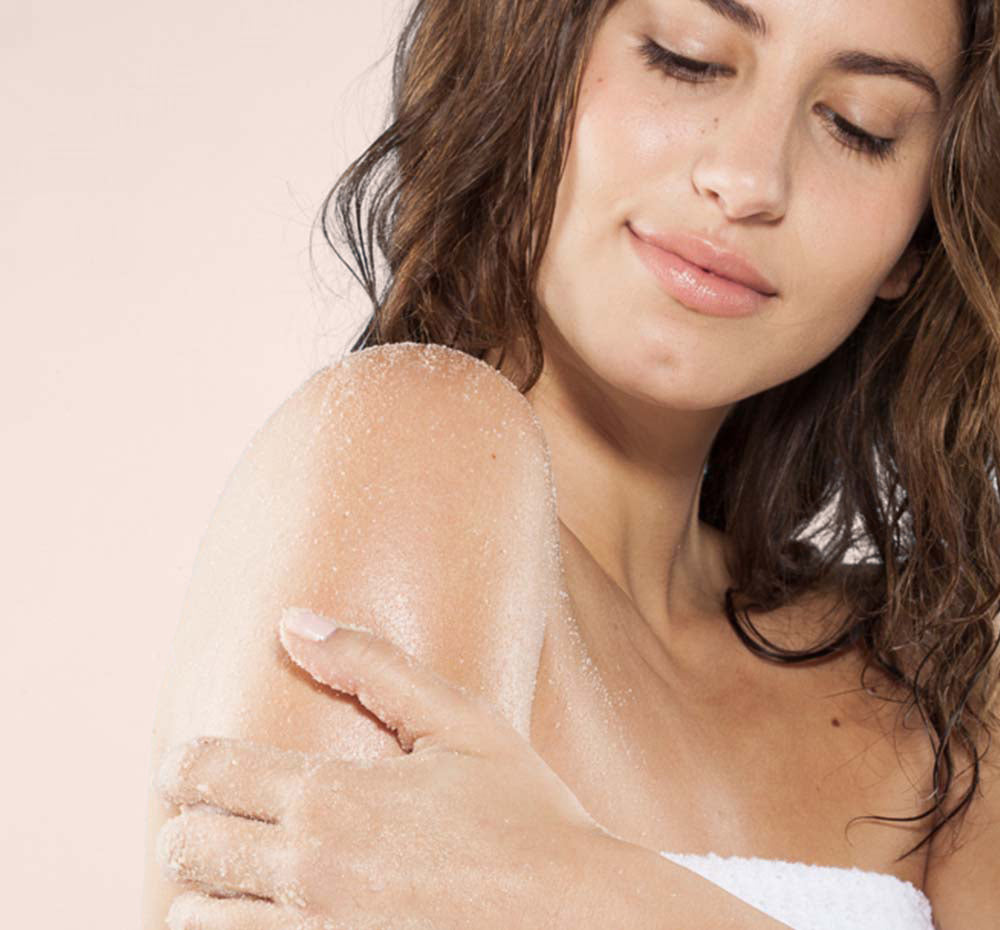 A woman is exfoliating her arm with FarmHouse Fresh Sweet Cream Fine Sea Salt Body Polish for soft and nourished skin.