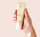 A hand holding a tube of Sweet Tea Shea Butter travel-size cream by FarmHouse Fresh scented with notes of peach, ginger, orchid and white tea.