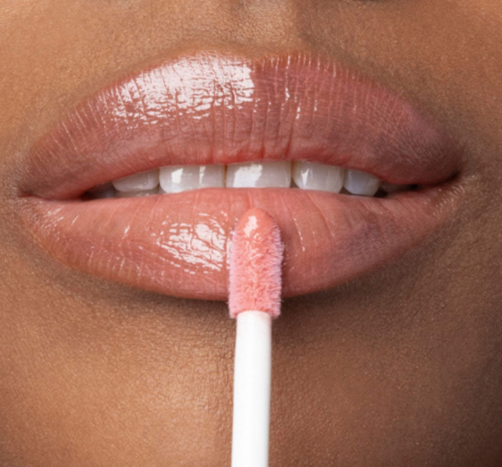 A woman is applying FarmHouse Fresh Vitamin Glaze Lip Gloss in Peach Peony color that restores dull  lips.