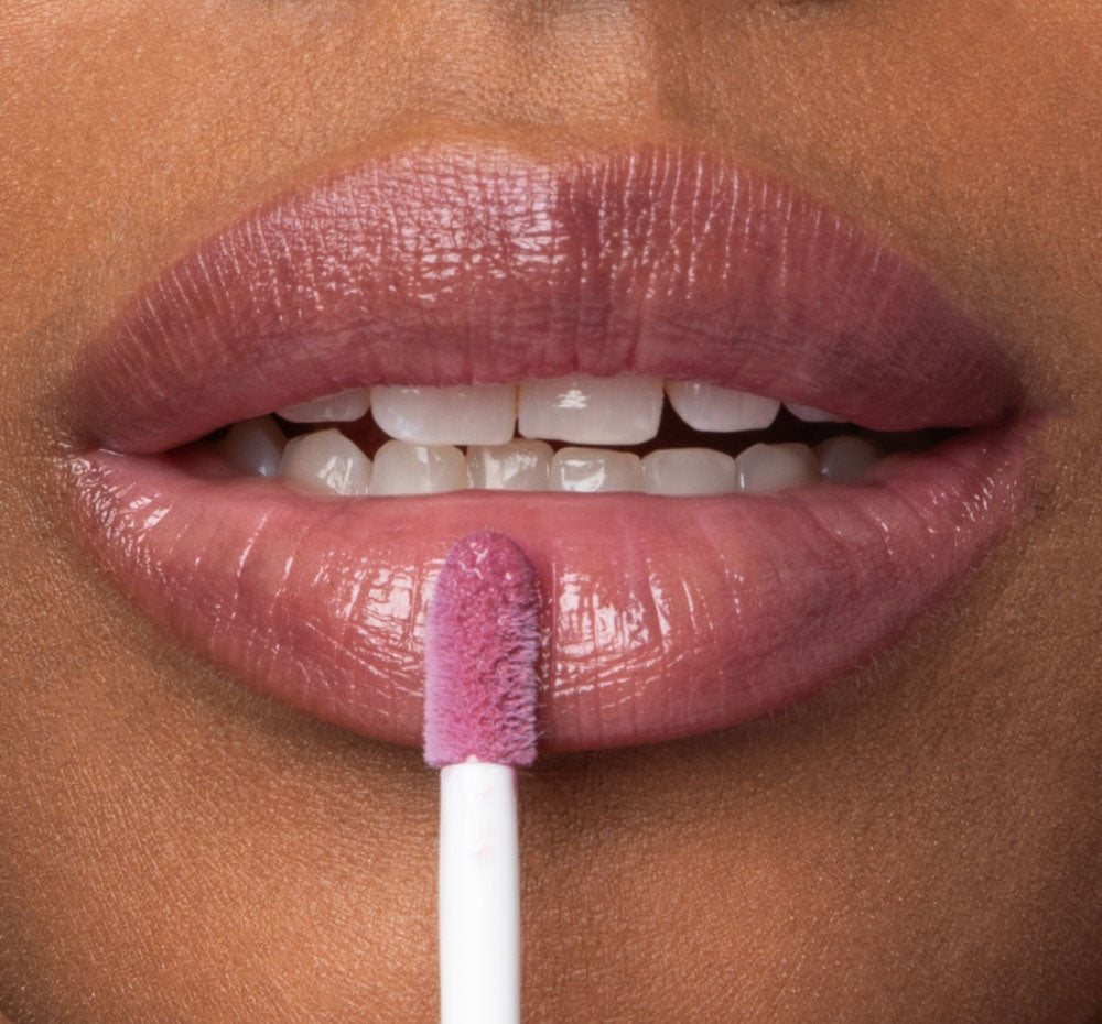 A woman is applying FarmHouse Fresh Vitamin Glaze Lip Gloss in Violet Orchid color that brings color to lips.