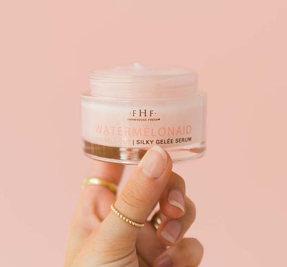 A person holding a jar of Farmhouse Fresh Watermelonaid cream infused with watermelon extract and CBD on a pink background.