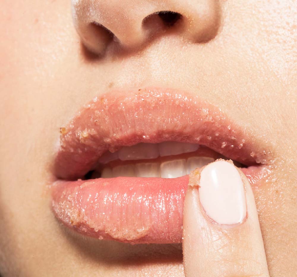 A close up of a woman exfoliating her lips with Farmhouse Fresh Beach Punch lip polish with a tropical vibe.