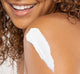 A woman with curly hair smiling while applying moisturizing Blissed Moon Dip weightless whip cream on her arm.