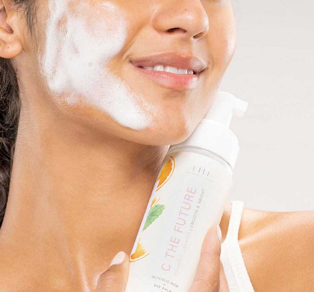 A woman is washing her face with FarmHouse Fresh C the Future Foam Facial Cleanser to exfoliate, smooth and brighten the look of skin.