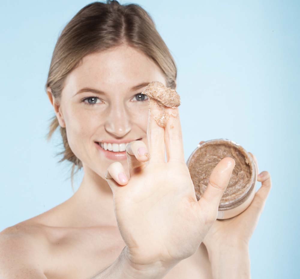 A woman is holding a jar of Farmhouse Fresh Coconut Beach exfoliating and moisturizing body scrub.