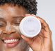 A woman with Pajama Paste Soothing Active Yogurt Mask by FarmHouse Fresh on her face and a jar in her hand, smiling while enjoying the pleasant honey scent of the product.