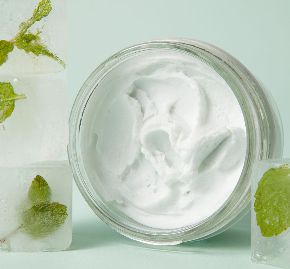 An opened of FarmHouse Fresh Enrich Mint Foot Rescue Mask next to ice cubes with mint leaves that represent the cooling effect of this leave-on rescue mask for feet that instantly brings comfort to dry skin.