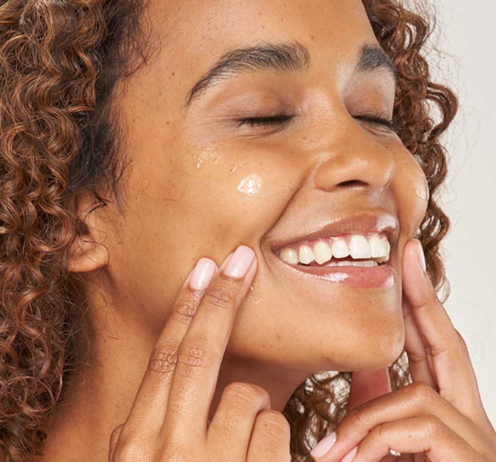 A woman is applying FarmHouse Fresh Green Fixer Calming Elixir Organic Matcha Face Serum on her face to hydrate and soothe skin.