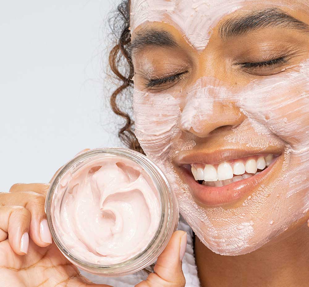 A woman with FarmHouse Fresh Mighty Brighty Vitamin C + Chamomile Brightening Mask on her face is holding a jar of the mask and smiling, enjoying the skin tightening effect.