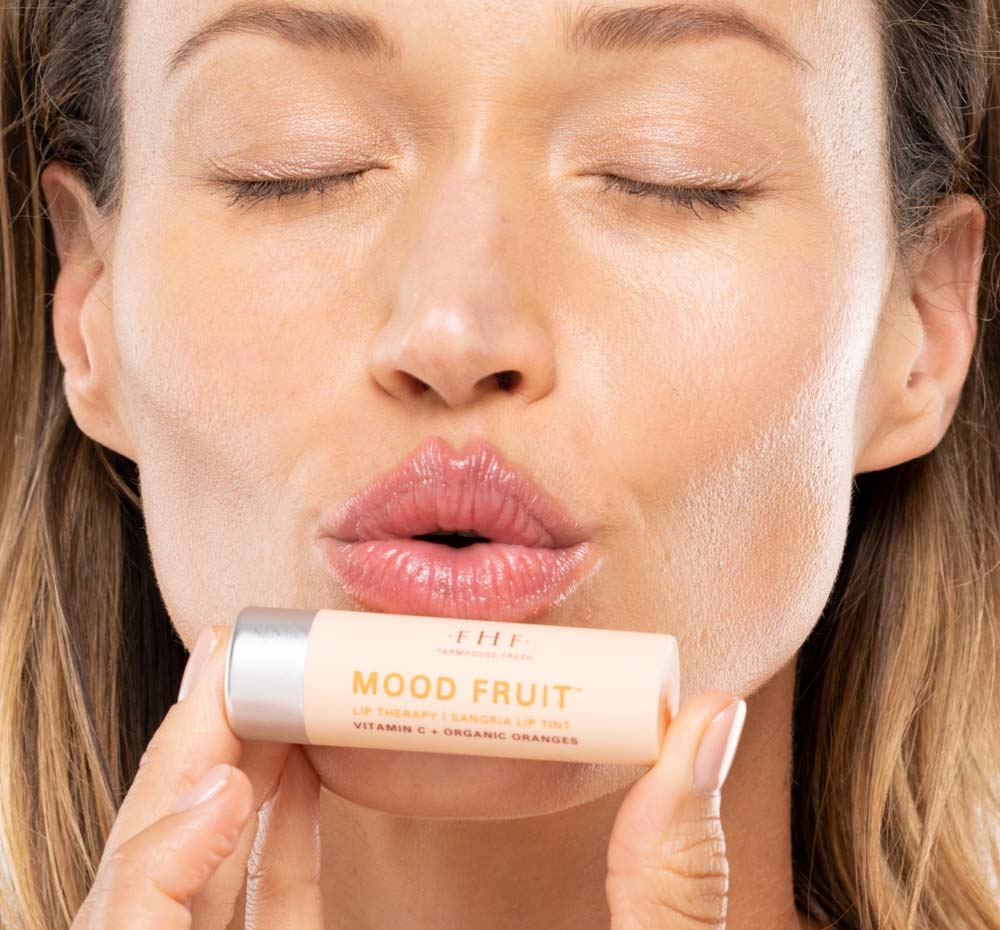 A woman is showing her hydrated and natural looking peachy lips after using FarmHouse Fresh Orange Mood Fruit Lip balm.