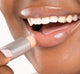 A woman is applying FarmHouse Fresh Orange Mood Fruit tinted lip balm to moisturize her lips with a slight touch of color.