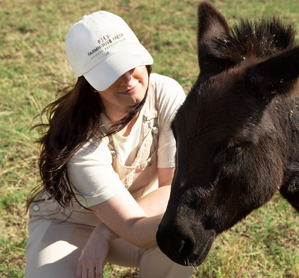 A woman wearing FarmHouse Fresh baseball hat with “Donkey Rescuer Club” embroidered on the front, sitting next to a donkey.