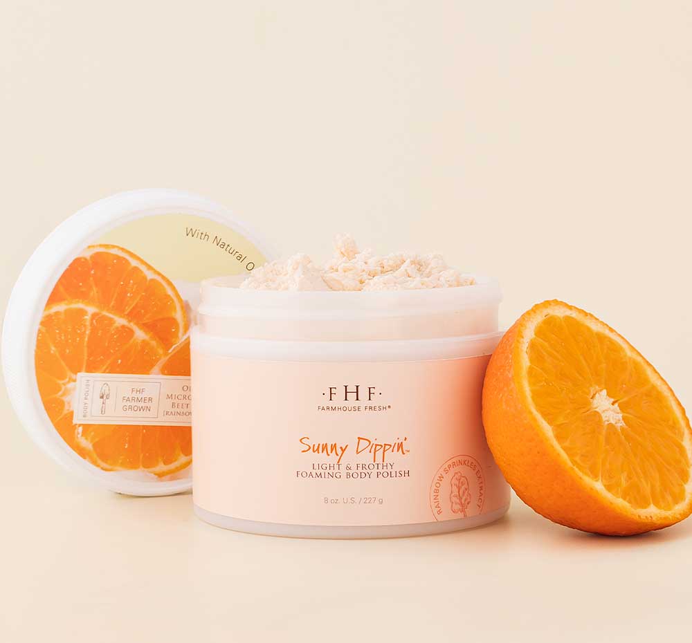 A jar of FarmHouse Fresh Sunny Dippin’ Foaming Body Polish that cleanses and buffs away rough skin, next to an orange.