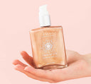 A hand holding a bottle of FarmHouse Fresh Sunshine Silk Shimmer Air Oil made with natural ingredients that moisturize and soften skin while giving it a rose-gold glow.