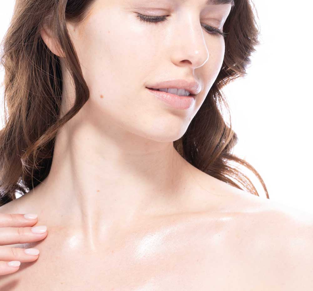 A woman is applying FarmHouse Fresh Sunshine Silk Shimmer Air Oil to her décolleté to achieve a rose-gold glow and soft skin. 