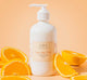 A bottle of FarmHouse Fresh Sunshine Silk Shea Butter body moisturizer scented with notes of creamy vanilla and juicy tangerines, next to orange slices.