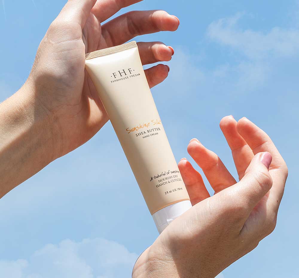 Hands holding a tube of rich, moisturizing Sunshine Silk shea butter cream by FarmHouse Fresh with the sky in the background.