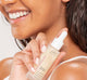 A smiling woman is holding a bottle FarmHouse Fresh Vitamin C Booster, demonstrating her bright, soft and smooth skin.
