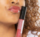 A woman holding FarmHouse Fresh Vitamin Glaze Oil Infused Lip Gloss in Berry color by her face, demonstrating the rich color of the lip gloss and her soft, well moisturized lips.