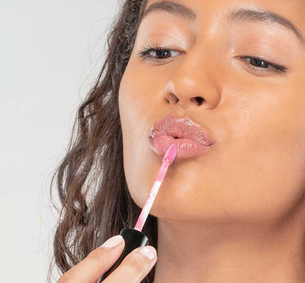 A woman is applying FarmHouse Fresh Vitamin Glaze Lip Gloss in Sheer Pink color that brings color and hydrates lips.