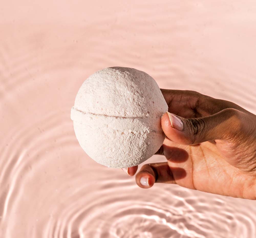 A hand holding FarmHouse Fresh Watermelon Basil Hemp-Infused Large Fizzing Bath Soak with Swellness Fortune above bath water.