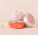 A jar of Farmhouse Fresh Watermelonaid cream infused with watermelon extract and CBD on top of a watermelon slice on a pink background.