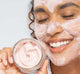 A woman with FarmHouse Fresh Mighty Brighty Vitamin C + Chamomile Brightening Mask on her face is holding a jar of the mask and smiling, enjoying the skin tightening effect.